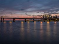 a bridge spanning over the ocean next to buildings at sunset on a pier with a moonlit sky and light reflection