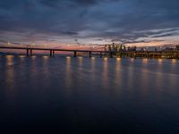 a bridge spanning over the ocean next to buildings at sunset on a pier with a moonlit sky and light reflection