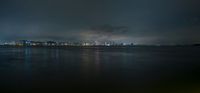 the water is dark and cloudy near the city skylines and lights are on at night