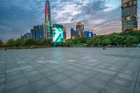 a woman is walking in the foreground of an empty city square at dusk with skyscrapers in the background