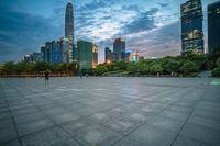 a woman is walking in the foreground of an empty city square at dusk with skyscrapers in the background