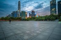 a woman is walking in the foreground of an empty city square at dusk with skyscrapers in the background