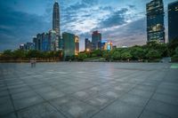 a woman is walking in the foreground of an empty city square at dusk with skyscrapers in the background