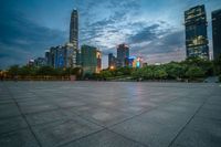 a woman is walking in the foreground of an empty city square at dusk with skyscrapers in the background