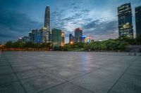 a woman is walking in the foreground of an empty city square at dusk with skyscrapers in the background