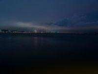 a lake with lights on the waterfront and a boat in the water at night on a cloudy day