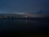 a lake with lights on the waterfront and a boat in the water at night on a cloudy day