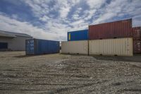 several shipping containers lined up in front of some building and sky behind them, on dirt and gravel