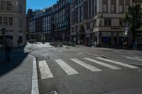 a cross walk is lined with buildings, on which people are riding bicycles, a pedestrian is walking on the sidewalk, and people on a bike are on the street