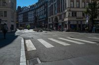 a cross walk is lined with buildings, on which people are riding bicycles, a pedestrian is walking on the sidewalk, and people on a bike are on the street