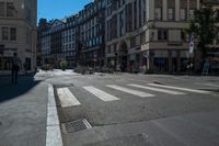 a cross walk is lined with buildings, on which people are riding bicycles, a pedestrian is walking on the sidewalk, and people on a bike are on the street