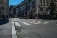 a cross walk is lined with buildings, on which people are riding bicycles, a pedestrian is walking on the sidewalk, and people on a bike are on the street