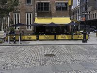 the sidewalk of a brick restaurant near two buildings with black and yellow striped tables in front