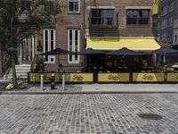 the sidewalk of a brick restaurant near two buildings with black and yellow striped tables in front