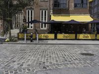 the sidewalk of a brick restaurant near two buildings with black and yellow striped tables in front