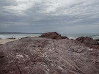 a person is sitting on the edge of a rocky ledge at the beach with the ocean behind him