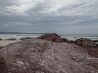 a person is sitting on the edge of a rocky ledge at the beach with the ocean behind him