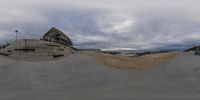 a very big skateboard park next to a beach, the sky and buildings are in a cloudgy sky