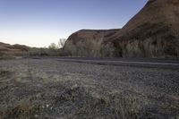 a skateboarder on a dirt road in the desert during sunset time, looking at a rocky hill and horizon