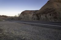 a skateboarder on a dirt road in the desert during sunset time, looking at a rocky hill and horizon