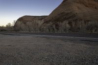 a skateboarder on a dirt road in the desert during sunset time, looking at a rocky hill and horizon