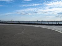 a woman is riding her skateboard by the water side of a pier with cars in the distance