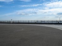 a woman is riding her skateboard by the water side of a pier with cars in the distance