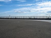 a woman is riding her skateboard by the water side of a pier with cars in the distance