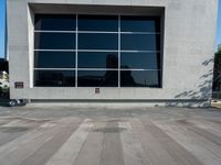 the skateboarder is riding on the concrete pavement by the wall with large windows