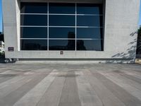 the skateboarder is riding on the concrete pavement by the wall with large windows