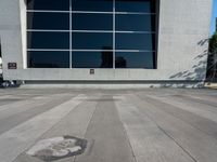 the skateboarder is riding on the concrete pavement by the wall with large windows