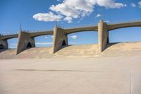 a skateboarder at the skate park in front of a bridge and stairs, against a cloudy blue sky