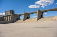 a skateboarder at the skate park in front of a bridge and stairs, against a cloudy blue sky