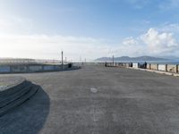 skateboarders are riding on a paved surface, near a fence and sea with a blue sky