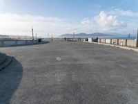 skateboarders are riding on a paved surface, near a fence and sea with a blue sky