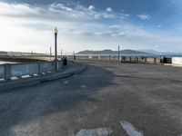 skateboarders are riding on a paved surface, near a fence and sea with a blue sky