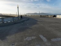 skateboarders are riding on a paved surface, near a fence and sea with a blue sky