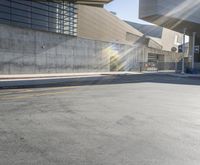 a skateboarder riding down a street in a parking lot near large buildings, with sun shining through the windows