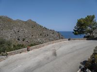 a person rides a skateboard down a rocky street next to the ocean and a mountain side