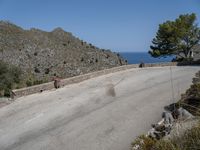 a person rides a skateboard down a rocky street next to the ocean and a mountain side