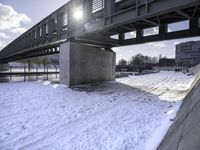 a man riding a skateboard across a snowy riverbed with the sun peeking through the bridge