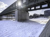 a man riding a skateboard across a snowy riverbed with the sun peeking through the bridge