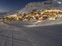 a ski slope going up a snow covered hillside with houses lit up at night in the background