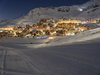 a ski slope going up a snow covered hillside with houses lit up at night in the background