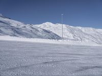 two people on a ski slope going down a snowy mountain side area in the foreground, with a tall snow covered hill behind them and a small lamp post in the top