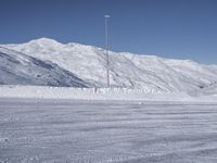 two people on a ski slope going down a snowy mountain side area in the foreground, with a tall snow covered hill behind them and a small lamp post in the top