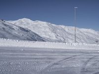 two people on a ski slope going down a snowy mountain side area in the foreground, with a tall snow covered hill behind them and a small lamp post in the top