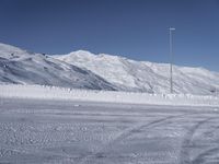 two people on a ski slope going down a snowy mountain side area in the foreground, with a tall snow covered hill behind them and a small lamp post in the top