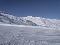 two people on a ski slope going down a snowy mountain side area in the foreground, with a tall snow covered hill behind them and a small lamp post in the top