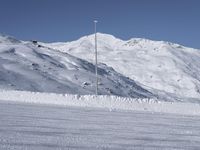 two people on a ski slope going down a snowy mountain side area in the foreground, with a tall snow covered hill behind them and a small lamp post in the top